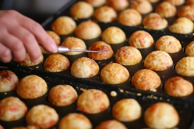 Hand turning golden takoyaki balls with a metal pick on a cast-iron griddle at a street food stall