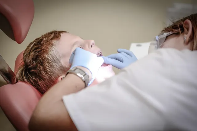 Dentist in blue gloves and safety glasses examining a young boy's teeth as he reclines in a red dental chair