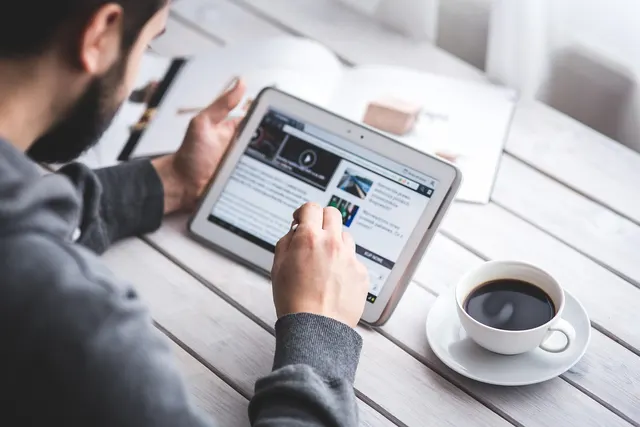 Bearded man in gray sweater browsing a website on a tablet at a white wooden table beside a cup of black coffee
