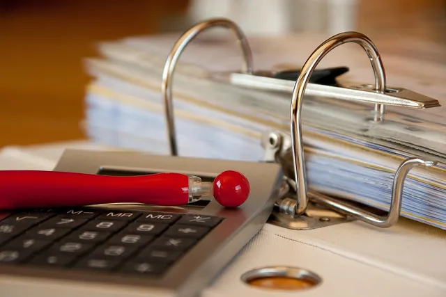 Calculator with a red pen in front of an open ring binder stuffed with paperwork on a desk