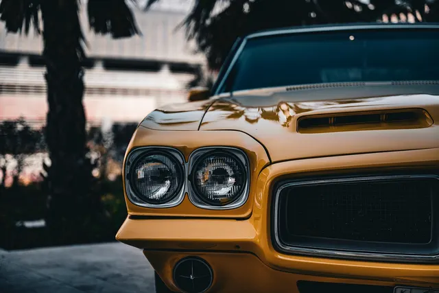 Close-up of a yellow vintage muscle car's front end with dual round headlights, hood scoop and grille, palm trees behind