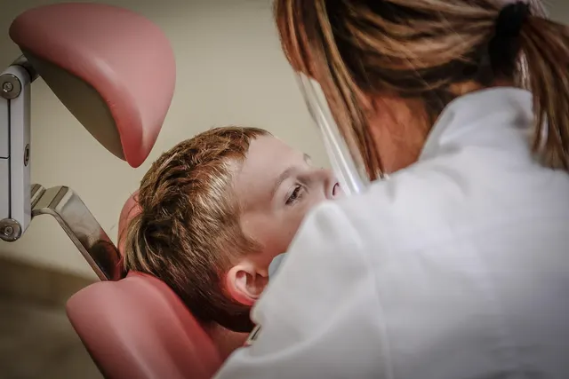 Young boy reclining in a pink dental chair while a dentist in a white coat and face shield examines him