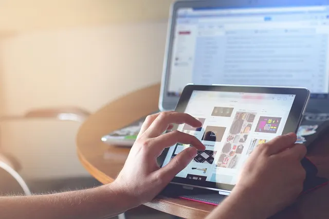 Hands browsing a product image grid on a tablet at a wooden table, with an open laptop showing an email inbox behind