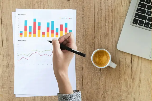 Hand marking a printed bar chart beside coffee and a laptop