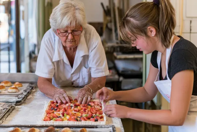 Older baker in glasses and a young woman in an apron arrange strawberries and fruit on a tart in a bakery kitchen