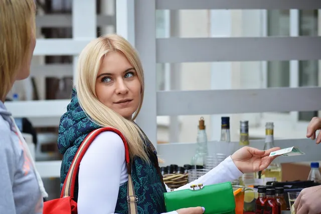 Blonde woman holding a green wallet hands cash to a vendor at an outdoor market stall with bottles on the counter
