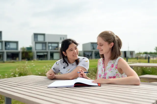 Two smiling young women at an outdoor wooden table reviewing an open notebook together, one holding a pen