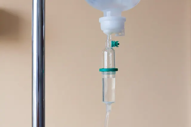 Close-up of an IV drip bag and clear drip chamber with tubing hanging beside a chrome IV pole against a beige wall