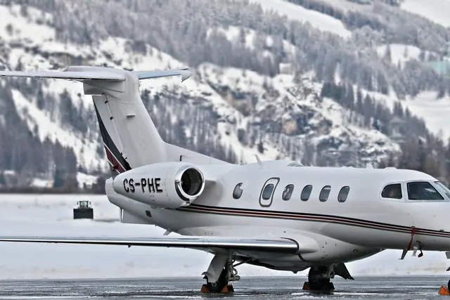 White private jet with red and black stripes parked on a snowy airport apron, snow-covered forested mountains behind