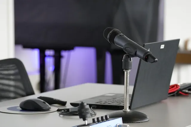 Desk with a handheld microphone on a stand, open laptop, mouse and audio interface in a purple-lit recording space