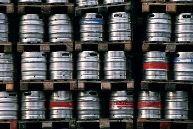 Rows of stainless steel beer kegs, some with red and blue bands, stacked on wooden pallets in a warehouse