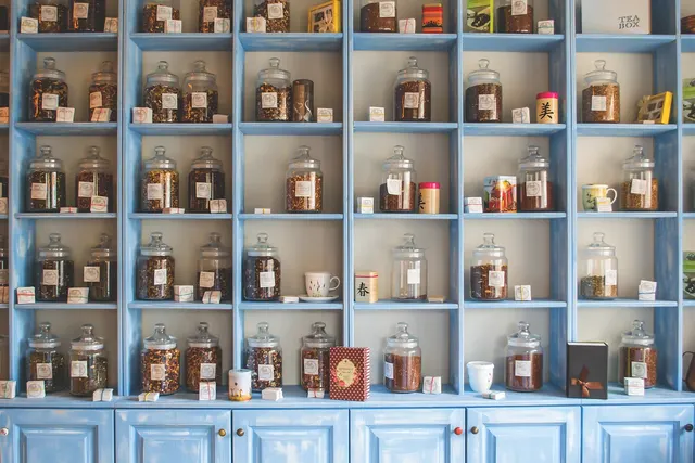 Blue wooden shelves in a botanical shop lined with labeled glass jars of loose dried herbs and tea, plus small tins and cups