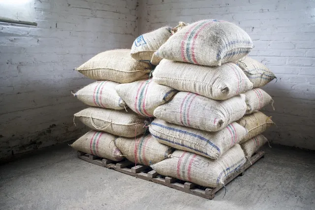 Burlap sacks with red and blue stripes stacked on a wooden pallet in a white brick storeroom, ready for bulk shipment