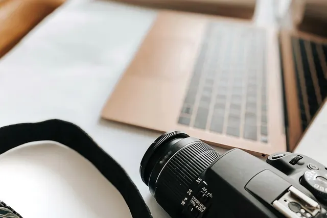 DSLR camera with neck strap in the foreground beside an open rose gold laptop on a white desk