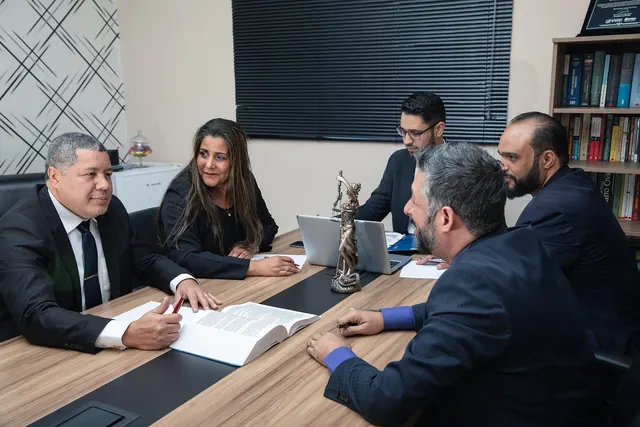 Five lawyers in suits meet at a conference table with an open law book, laptop and Lady Justice statue in an office