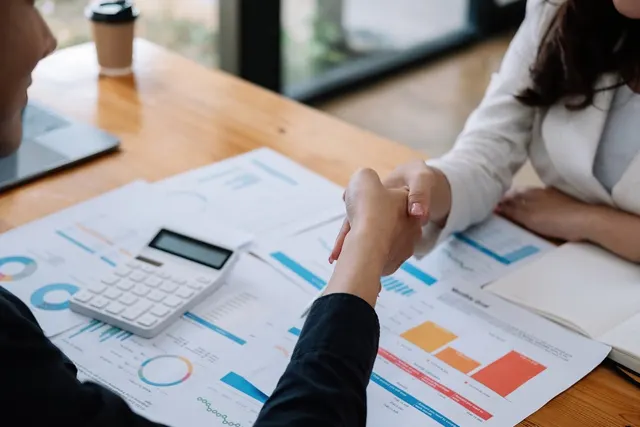 Two professionals shake hands across a wooden desk covered with financial charts, a calculator and a laptop