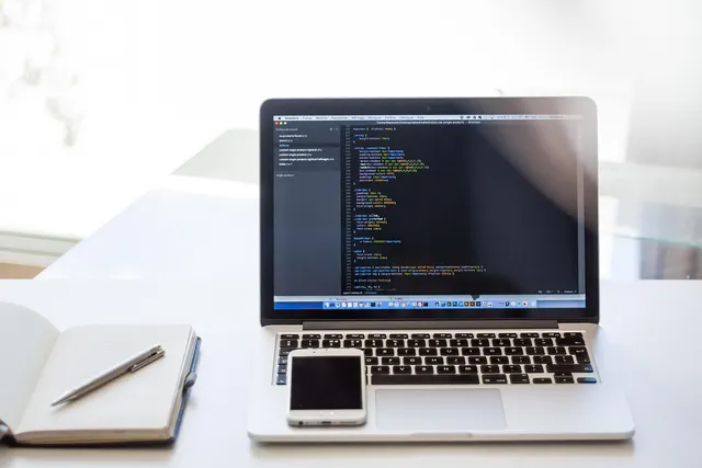Laptop showing code in an editor with a smartphone on its trackpad beside an open notebook and pen on a white desk