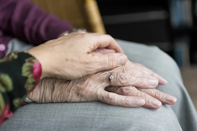 Caregiver's hand resting on an elderly woman's wrinkled hands, which wear a diamond ring, folded on her gray-trousered lap