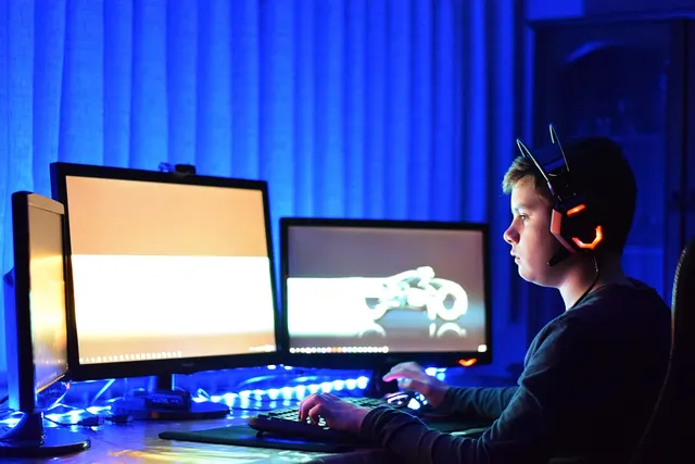 Boy wearing a glowing gaming headset plays at a desk with three monitors, keyboard and mouse in a blue-lit room