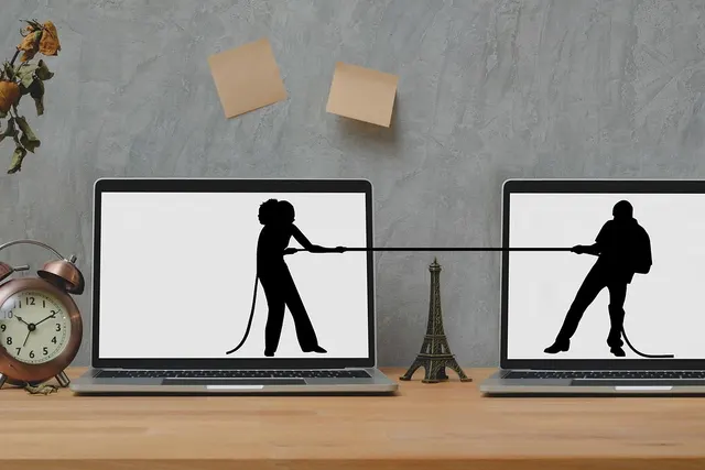 Two laptops on a wooden desk showing silhouettes in a tug-of-war, beside an alarm clock and a vase of dried roses
