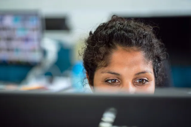 Woman with curly dark hair peers intently over a computer monitor in an office, blurred screens behind her