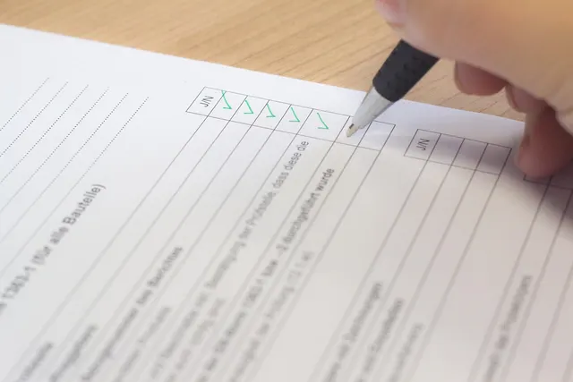 Hand holding a pen ticking green checkmarks in yes/no boxes on a printed compliance checklist form