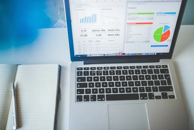Laptop on a white desk showing a financial dashboard with bar and pie charts, beside an open notebook and silver pen