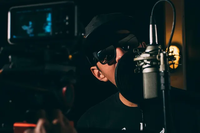Content creator in a cap and sunglasses speaking into a studio microphone with pop filter as a camera films him