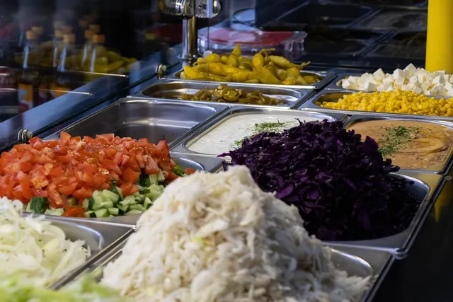Restaurant prep counter with steel trays of shredded cabbage, red cabbage, diced tomatoes, cucumber, peppers, corn and feta