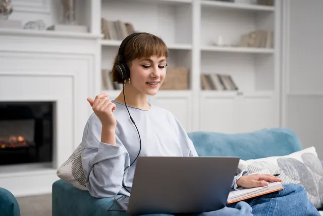 Smiling woman in headphones using a laptop on a blue sofa at home, holding a pen over a notebook