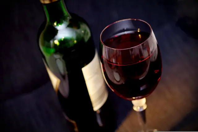Glass of red wine beside a dark green wine bottle with a plain cream label against a dark background