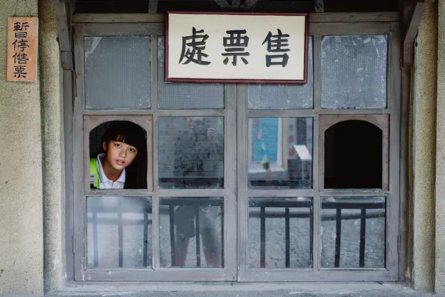 Girl peering out of an arched opening in a weathered wooden ticket booth window under a Chinese ticket office sign