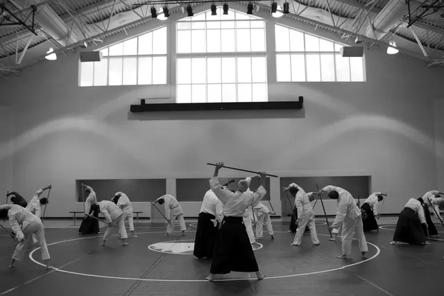 Black-and-white photo of aikido students in gi and hakama stretching with wooden staffs in a bright gym dojo