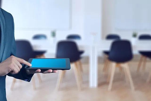 Professional in a blue suit tapping a tablet beside an empty conference room with dark chairs and white tables
