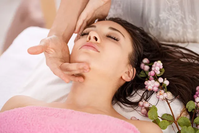 Woman with closed eyes receiving a facial massage on a spa table, wrapped in a pink towel beside pink berry sprigs