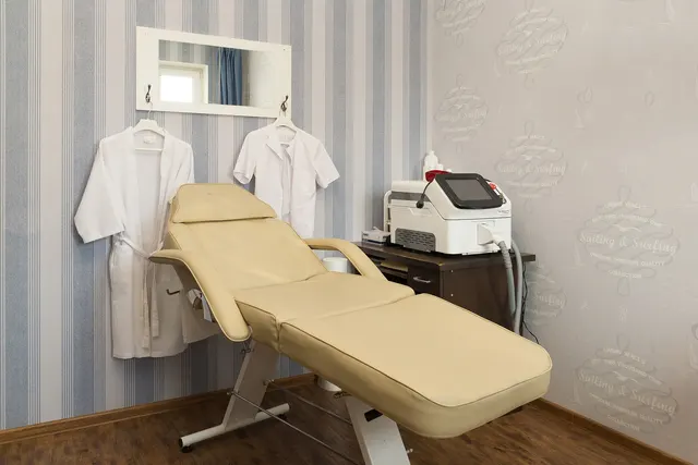 Med spa treatment room with a beige reclining treatment chair, a laser device on a cabinet and white robes on hooks