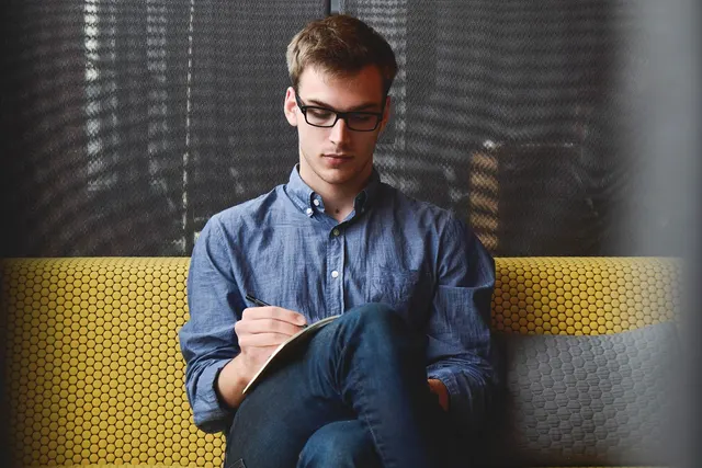 Young man in glasses and blue button-down shirt writing notes in a notebook while seated on a yellow couch