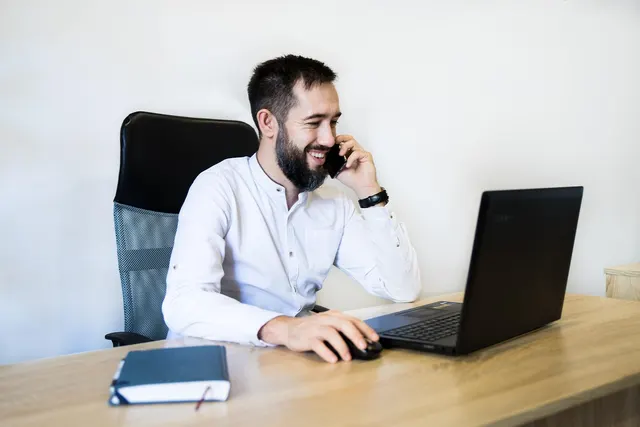 Smiling bearded man in a white shirt talks on his phone while using a laptop at a wooden office desk