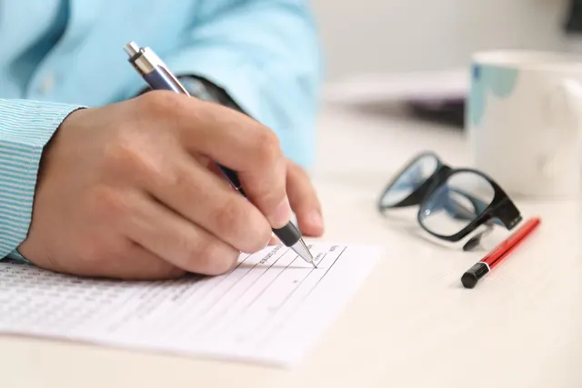 Person in a light blue striped shirt filling out a paper form with a pen at a desk beside glasses and a mug