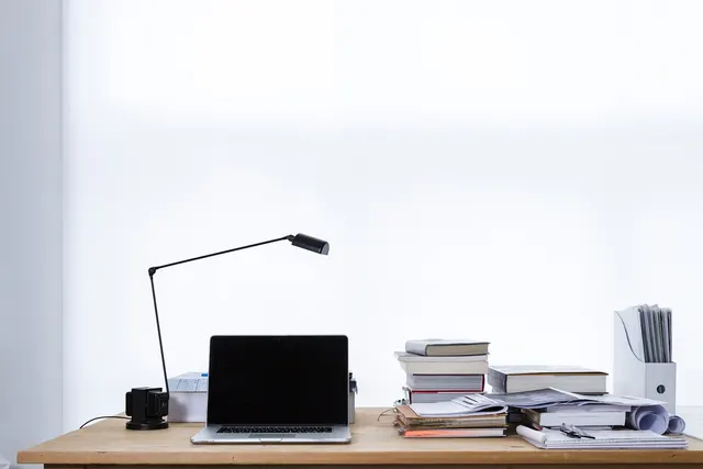 Wooden desk with an open laptop, black desk lamp, stacked books and papers, and a magazine file holder against a white wall