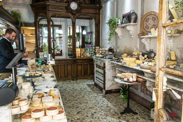 Cheese shop interior with a man at the counter by a card terminal, cheese-filled marble shelves and a mosaic floor