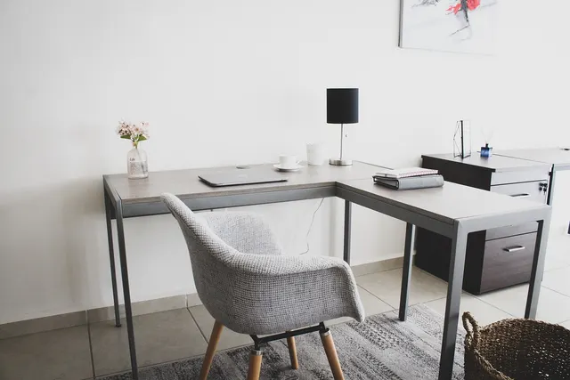 Minimal home office with an L-shaped desk, closed laptop, black lamp, coffee cup, binders and a grey upholstered chair