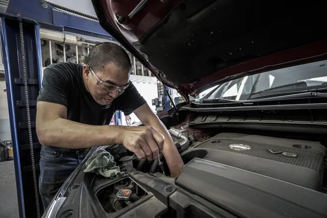 Mechanic in glasses and a black T-shirt leaning under a raised hood, wrenching on a red car's engine with a rag in hand