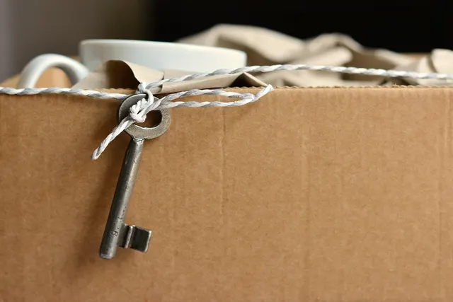 Vintage skeleton key tied with twine to a cardboard moving box packed with a white mug and brown wrapping paper