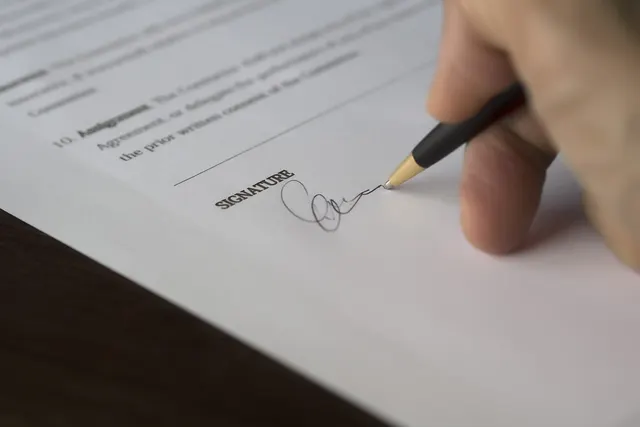 Close-up of a hand signing a printed contract with a black and gold pen on the signature line at a dark wooden desk