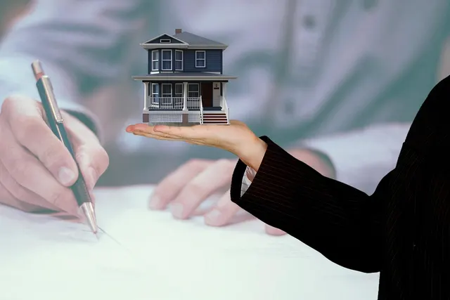 Hand in a pinstripe suit holding a miniature blue house over a faded image of a person signing a loan document