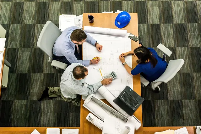Overhead view of three professionals at a table reviewing blueprints with a calculator, laptop and blue hard hat
