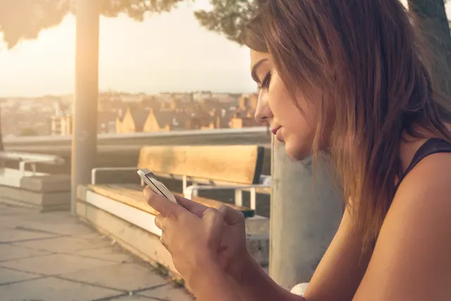 Young woman with long hair looking at her smartphone on a city terrace at sunset, wooden bench behind her