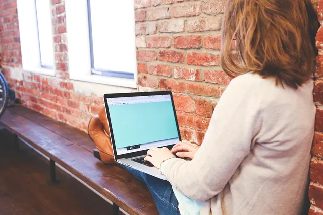 Woman with auburn hair typing on a laptop while seated on a wooden bench against a red brick wall