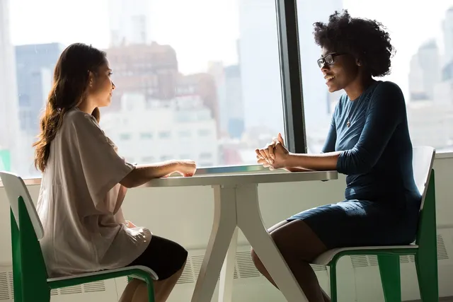 Two women talking across a white table by a large window overlooking city skyscrapers in a bright office